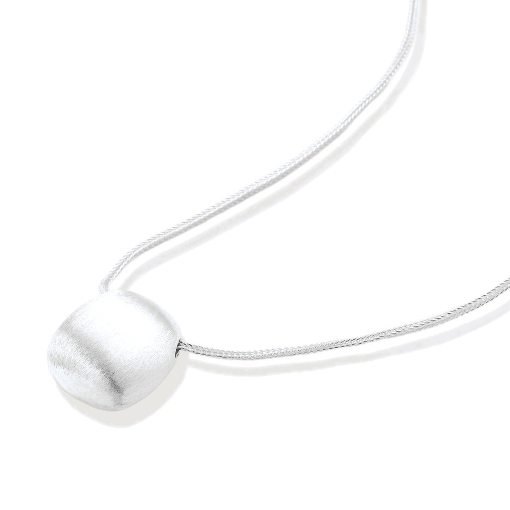 Minimalist brushed sterling silver dome pendant on delicate chain against cream background, showing matte textured finish