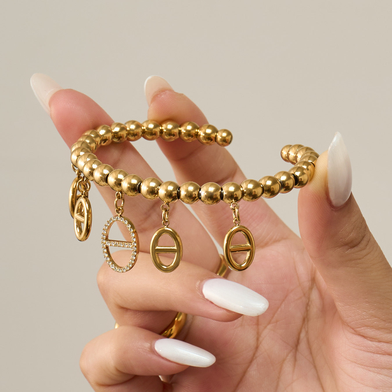 Gold bracelet with charms held by a hand with white nail polish on a neutral background