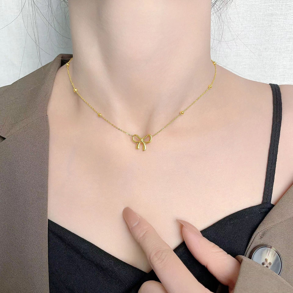 Gold necklace with a bow pendant worn by a person, pointing to the necklace.
