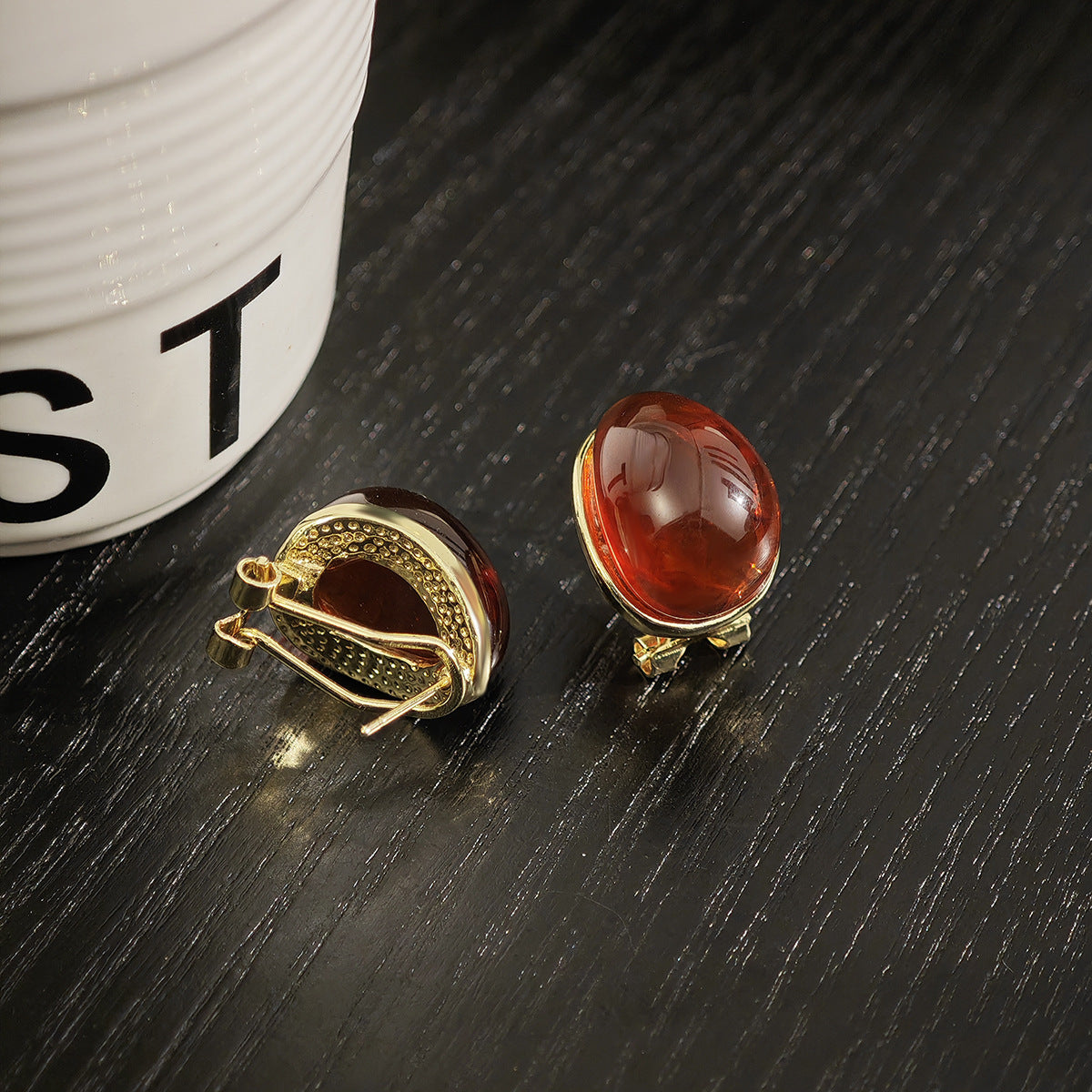 Two amber-colored gemstone earrings on a dark surface with a white container in the background.