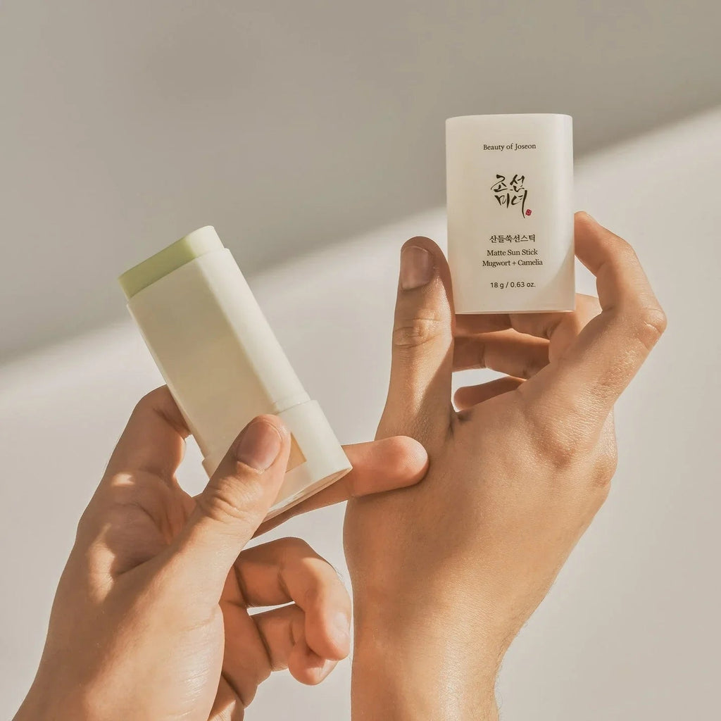 Two hands holding a white cosmetic stick and a small container with text on a light background
