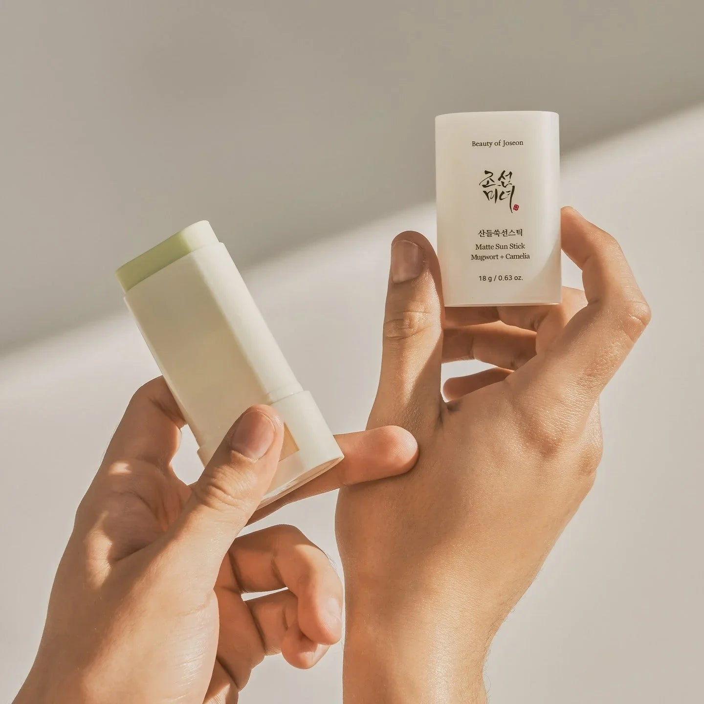 Two hands holding a white cosmetic stick and a small container with text on a light background